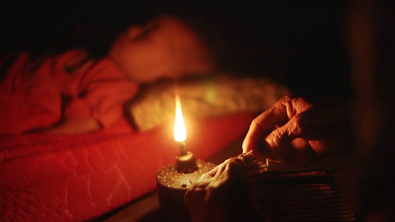 写真：灯油ランプのあかりのもとで縫物をする老人の手元。あかりの向こうには赤ちゃんが眠っている